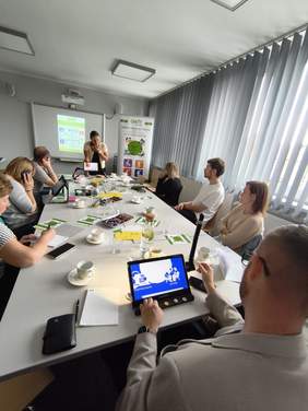 Pokaż zdjęcie:  Uczestnicy szkolenia siedzą przy długim stole, na którym leżą dokumenty i kubki. Większość osób patrzy na ekran projekcyjny z jasnym slajdem na przedniej ścianie. Obok ekranu stoi roll-up, a po prawej stronie znajdują się duże okna z pionowymi żaluzjami. Na pierwszym planie mężczyzna w jasnej marynarce trzyma tablet z niebieskim ekranem.