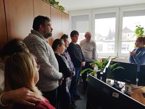 Pokaż zdjęcie: Grupa około ośmiu osób zebrana w półokręgu w biurze. Po prawej stronie kobieta w niebieskiej bluzie zwraca się do pozostałych uczestników. W tle znajdują się duże okna z widokiem na sąsiednie budynki, a po lewej stronie wysoka, drewniana szafa. Na pierwszym planie widoczne są biurka z monitorami komputerowymi.