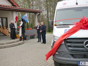 Pokaż zdjęcie: Na pierwszym planie widać przód białego samochodu przewiązanego czerwoną wstęgą. W tle mężczyźni, w tym dwóch księży, stoją na tle budynku. O budynek oparta jest rzeźba przedstawiająca trzy anioły.