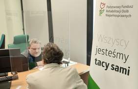 Pokaż zdjęcie: Mężczyzna siedzi przy biurku z komputerem. Przed nim kobieta w marynarce, która zadaje pytanie.  Po prawej stronie baner promujący PFRON.