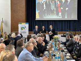 Pokaż zdjęcie: Kobieta ubrana elegancko trzyma mikrofon i mówi od wielu osób siedzących przy stołach ustawionych w rzędach. Po obu jej stronach stoją mężczyźni.