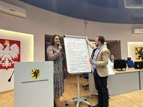 Pokaż zdjęcie: Kobieta w długiej sukni w kwieciste wzory trzyma mikrofon. Obok stoi mężczyzna w marynarce i koszuli poprawia duże kartki wiszące na tablicy. Po lewej stronie mównica i godło