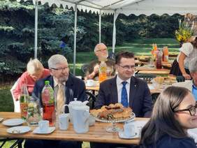 Pokaż zdjęcie: Przy stołach siedzą ludzie. Uśmiechają się, rozmawiają i jedzą. Na stołach stoją różna ciasta, kawa i napoje. 