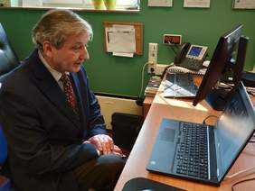 Pokaż zdjęcie: Fragment pomieszczenia mężczyzna w garniturze siedzi w fotelu przed sobą ma otwarty laptop który leży na biurku