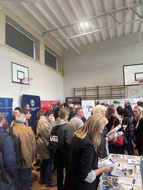 Pokaż zdjęcie: Tłum osób zgromadzonych przy stoiskach w sali gimnastycznej; uczestnicy czytają ulotki i rozmawiają, w tle widoczne banery informacyjne i kosz do koszykówki.