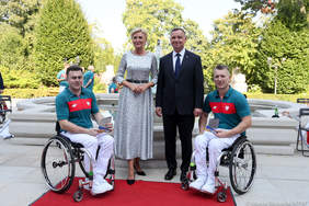 Pokaż zdjęcie: Para prezydencka stoi na tle fontanny znajdującej się w parku. Zarówno z prawej i lewej ich strony znajdują się poruszający się na wózkach sportowcy.