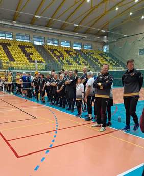 Pokaż zdjęcie: Ujęcie przedstawiające dwie drużyny Piłki Siatkowej na Siedząco stojące w rzędach naprzeciwko siebie wzdłuż siatki, prawdopodobnie podczas ceremonii otwarcia lub zamknięcia turnieju. Zawodnicy ubrani są w ciemne, sportowe stroje. Wyróżniają się trenerzy lub członkowie sztabu, którzy mają na sobie czarno-żółte dresy, a jeden z mężczyzn po prawej ma wyraźnie widoczną brodę i koszulkę z logo w kolorze złoto-czarnym. W grupie widać także osoby z widocznymi protezami lub kulami. Hala sportowa ma żółte trybuny w tle i wysoki, łukowaty sufit. Na pierwszym planie widać wyraźnie linie boiska.