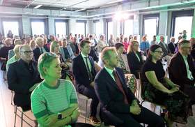 Publiczność siedząca w rzędach w przestronnej sali konferencyjnej z wysokim sufitem podczas finału ogólnopolskiego konkursu architektonicznego. Na pierwszym planie od lewej siedzą: kobieta w jasnozielonej bluzce w kratkę, mężczyzna w ciemnym garniturze z czerwonym krawatem, kobieta w czarnej bluzce oraz mężczyzna w garniturze bez krawata. Wśród zaproszonych gości jest Prezes Zarządu PFRON Małgorzata Lorek oraz dyrektor Oddziału Wielkopolskiego PFRON Anna Skupień. Pomieszczenie oświetla wpadające przez duże okna dzienne światło o lekkim, zielonkawym odcieniu.