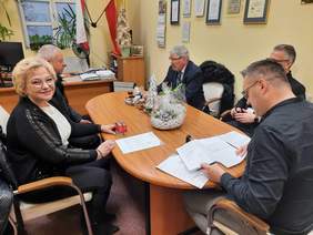 Pokaż zdjęcie: Przy stole siedzi pięć osób: jedna kobieta i czterech mężczyzn. Rozmawiają ze sobą. Na stole leżą różne dokumenty, pieczątki, stroiki świąteczne. 