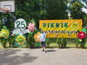 Pokaż zdjęcie: Stojący mężczyzna na wybrukowanym placu przed plenerową scenografią jubileuszową. Za mężczyzną w centrum stoi żółty baner z napisem „Piknik integracyjny” i logo „Warsztatu Terapii Zajęciowej w Szamotułach”, a obok biały okrąg z napisem „25 lat WTZ”. Całość dekorują duże, kolorowe kwiaty, krzewy w donicach oraz wysokie drzewa i kosz do koszykówki w tle.