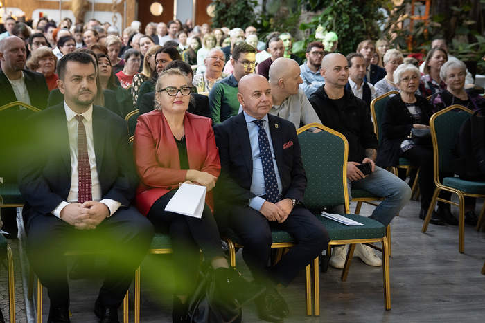 Pokaż zdjęcie: Na pierwszym planie Dyrektor Oddziału Lubuskiego PFRON wraz z Wiceprezydentem Zieonej Góry, a także inni uczestnicy spotkania, którzy są ubrali na galowo i siedzą na krzesłach, a w tle widoczne są rośliny w palmiarni