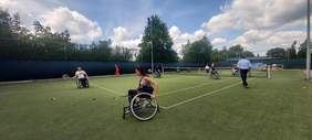 Pokaż zdjęcie: Trawiasty kort tenisowy na którym kilkanaście osób chodzących i poruszających się na wózkach inwalidzkich ćwiczy grę w tenisa.