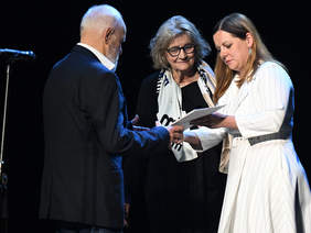 Pokaż zdjęcie: Trzy osoby na scenie w trakcie oficjalnej chwili. Kobieta w białej sukni wręcza dokument starszemu mężczyźnie. Obok nich stoi starsza kobieta w okularach, z szalem z napisem.