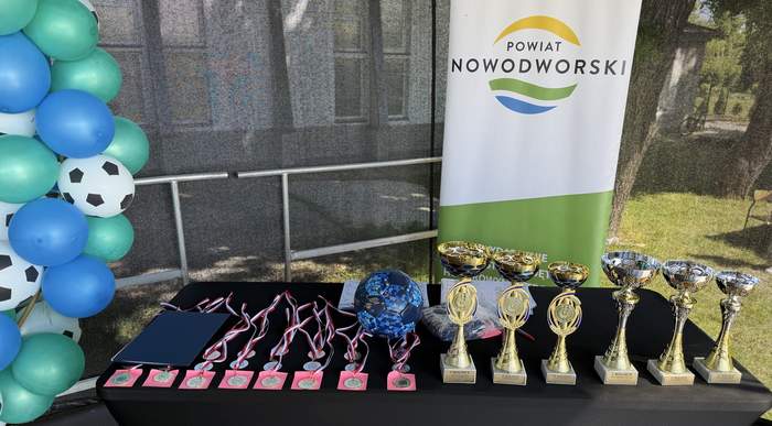 Pokaż zdjęcie: Stół z medalami, pucharami i nagrodami przygotowanymi dla uczestników wydarzenia sportowego. W tle widoczny baner Powiatu Nowodworskiego oraz dekoracja z niebiesko-zielonych balonów z motywem piłek.