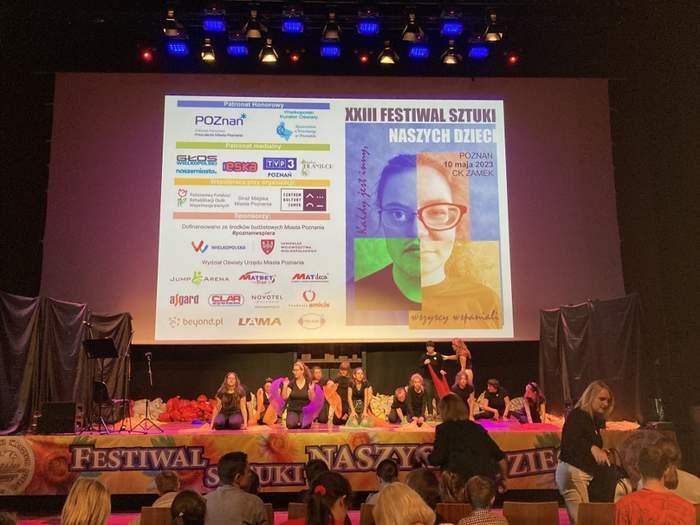Pokaż zdjęcie: Grupa młodych osób występuje na scenie teatralnej. Nad sceną, na dużym ekranie wyświetlony został plakat promujący XXIII Festiwal Sztuki Naszych Dzieci. Przed sceną zawieszony jest także baner promujący Festiwal. Możemy również dostrzec grupę osób, która siedzi na widowni.
