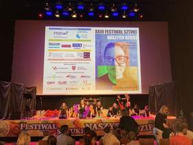 Pokaż zdjęcie: Grupa młodych osób występuje na scenie teatralnej. Nad sceną, na dużym ekranie wyświetlony został plakat promujący XXIII Festiwal Sztuki Naszych Dzieci. Przed sceną zawieszony jest także baner promujący Festiwal. Możemy również dostrzec grupę osób, która siedzi na widowni.