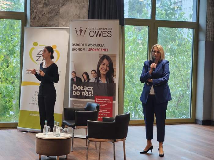 Pokaż zdjęcie: Po prawej stronie, na tle dużego okna, za którym można dostrzec dużo zieleni, stoi kobieta z mikrofonem. Po lewej stronie stoi druga kobieta, która tłumaczy spotkanie na język migowy, a w tle znajdują się dwa banery organizatorów konferencji.