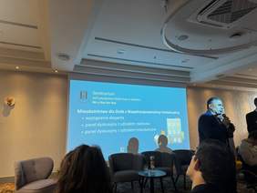 Pokaż zdjęcie: Prelegent przemawia podczas seminarium PSONI w Gdańsku na temat mieszkalnictwa dla osób z niepełnosprawnością intelektualną. Na niebieskim slajdzie widnieje program spotkania. Widownia siedzi w fotelach w nowoczesnej, jasnej sali konferencyjnej.