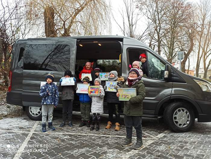 Pokaż zdjęcie: Dziesięcioro przedszkolaków w zimowych kurtkach i czapkach znajduje się przed lub w nowo zakupionym busie w rękach trzymają oprawione w srebrne ramki kolorowe rysunki pojazdu