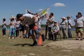 Pokaż zdjęcie: Jedna z konkurencji, młody zawodnik biegnie pochylony między pachołkami, kładzie na ziemi worki z piaskiem, w tle stoi duża grupa osób.