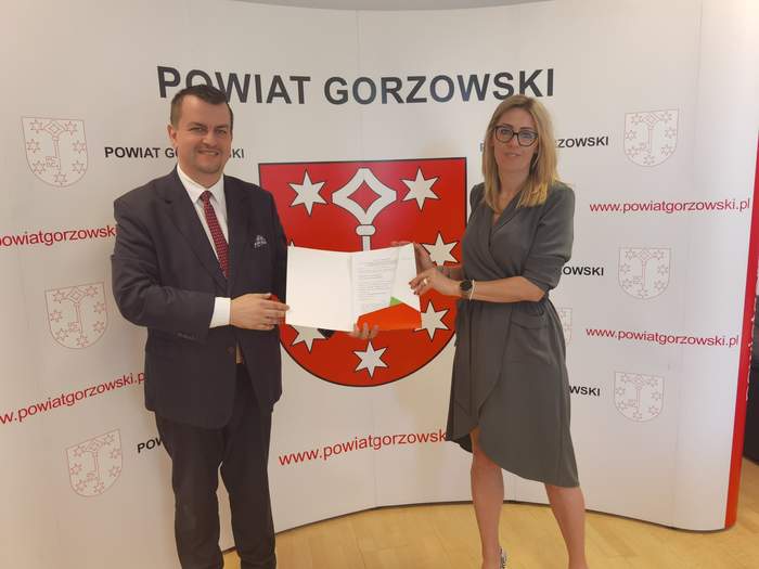 Pokaż zdjęcie: Dyrektor Oddziału Andrzej Gonia wraz ze Starostą Gorzowskim Magdaleną Pędziwiatr na tle herbu Powiatu Gorzowskiego z podpisaną umową