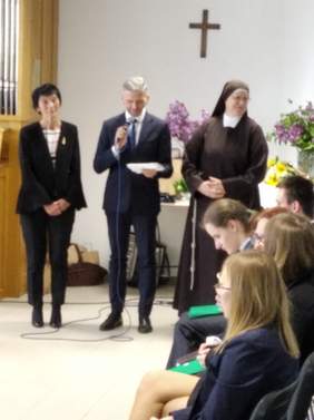 Pokaż zdjęcie: trzy osoby stoją. Jedna z nich mówi do mikrofonu. Na pierwszym planie cześć publiczności