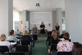 Pokaż zdjęcie: Cztery kobiety stoją zwrócono do widowni. Za nimi ekran z prezentacją Kompleksowej Rehabilitacji 