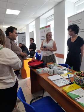 Pokaż zdjęcie: Na stoliku leżą ulotki, przy stoliku stoją osoby, które rozmawiają. W tle banery z logo PFRON, OWIT.