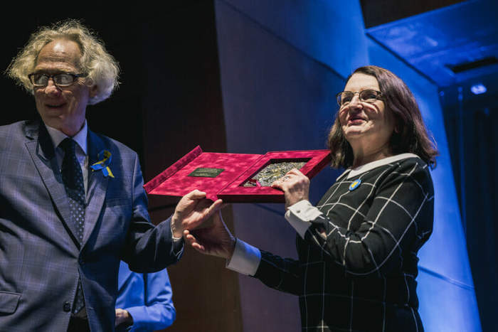 Pokaż zdjęcie: Mężczyzna w granatowym garniturze w kratkę , bląd włosach i okularach przekazuje pamiątką – grawer kobiecie w ciemnej marynarce w okularach i ciemnych dłuższych włosach. Obydwoje pozują do zdjęcia i się uśiechają.