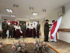 Pokaż zdjęcie: Na scenie WTZ Zgromadzenia Sióstr Służek NMPN w Szumowie stoi 9 osób: pięć kobiet i czterech mężczyzn. Mężczyźni przebrani są w mundury żołnierzy polskich, kobiety mają założone białe bluzki i bordowe, długie spódnice. Po prawej stronie, przy ścianie stoi kobieta ubrana w biało – czerwoną szatę oraz mężczyzna w mundurze wojskowym, który macha polską flagą.  