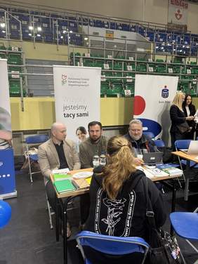 Pokaż zdjęcie: Na zdjęciu widać stoisko podczas targów pracy w Elblągu. Przy stole siedzą pracownicy firmy lub instytucji i rozmawiają z uczestniczką targów siedzącą tyłem do aparatu. Na stole leżą materiały informacyjne i napoje, a w tle znajdują się banery promocyjne oraz trybuny hali sportowej. Zdjęcie przedstawia moment rozmowy i prezentacji oferty odwiedzającym.