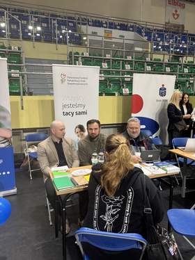 Na zdjęciu widać stoisko podczas targów pracy w Elblągu. Przy stole siedzą pracownicy firmy lub instytucji i rozmawiają z uczestniczką targów siedzącą tyłem do aparatu. Na stole leżą materiały informacyjne i napoje, a w tle znajdują się banery promocyjne oraz trybuny hali sportowej. Zdjęcie przedstawia moment rozmowy i prezentacji oferty odwiedzającym.