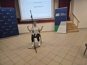 Kobieta w białej bluzce, czarnych getrach z opaską na głowie, siedzi, pokazuje na ramiona