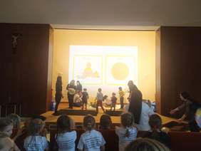 Pokaż zdjęcie: Na scenie leżą trzy dywany, siedzą lub stoją na nich przedszkolaki, na zdjęciu jest ogólne wrażenie zabawy. Z boku stoją opiekunowie dzieci, którzy pomagają w zabawie. Na ścianie wyświetlany jest piktogram z napisem bawimy się oraz żółtą piłką