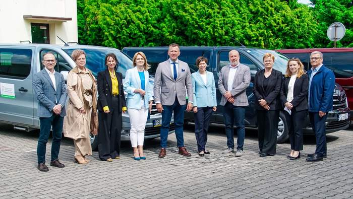 Pokaż zdjęcie: Grupa dziesięciu osób, kobiet i mężczyzn, stoi na chodniku, na tle trzech mikrobusów. Na busach widnieje naklejka informująca, że zostały dofinansowane ze środków PFRON. Z tyłu widoczny jest fragment budynku oraz wysoki żywopłot.