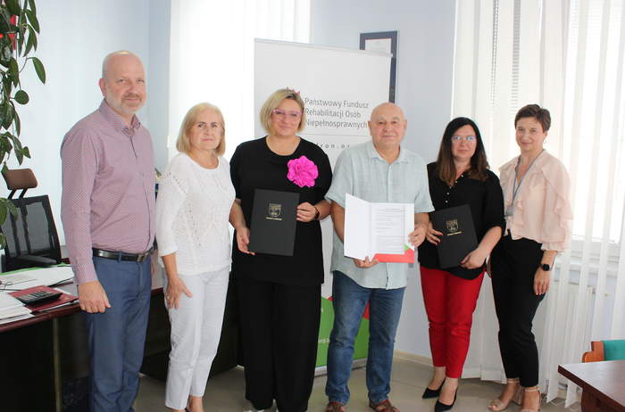 Pokaż zdjęcie: Pamiątkowe zdjęcie po podpisaniu umowy, na tle roollapu PFRON pozuje sześć osób. Dwie z nich trzymają w ręku umowy.