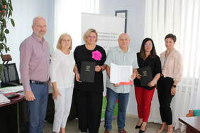 Pokaż zdjęcie: Pamiątkowe zdjęcie po podpisaniu umowy, na tle roollapu PFRON pozuje sześć osób. Dwie z nich trzymają w ręku umowy.