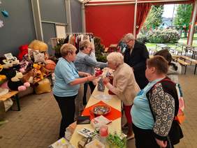 Pokaż zdjęcie: Grupa osób stoi przy kiermaszu, na którym można zakupić los. W tle widać różne maskotki, ubrania, rękodzieło i inne wyroby. 