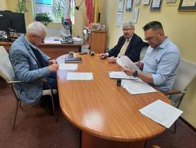 Pokaż zdjęcie: Przy podłużnym stole siedzi trzech mężczyzn. Podpisują umowę. Na stole leżą różne dokumenty, pieczątki, notesy i telefon. Na ścianach gabinetu wiszą różne dyplomy. 