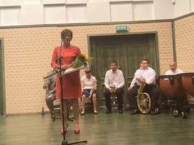 Pokaż zdjęcie: Przy mikrofonie stoi pani w pomarańczowej sukience, w tle orkiestra składająca się z osób niepełnosprawnych