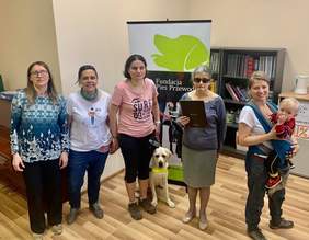 Pokaż zdjęcie: Na tle roll-up'u z napisem "Fundacja Pies Przewodnik" stoi pięć kobiet. Jedna kobieta trzyma małe dziecko na rękach. Pomiędzy nimi na podłodze leży pies rasy labrador w kolorze biszkopt.