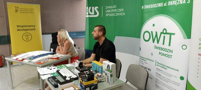 Pokaż zdjęcie: Kobieta i mężczyzna siedzą przy stolikach z materiałami informacyjnami, a za nimi rollupy z logo instytucji