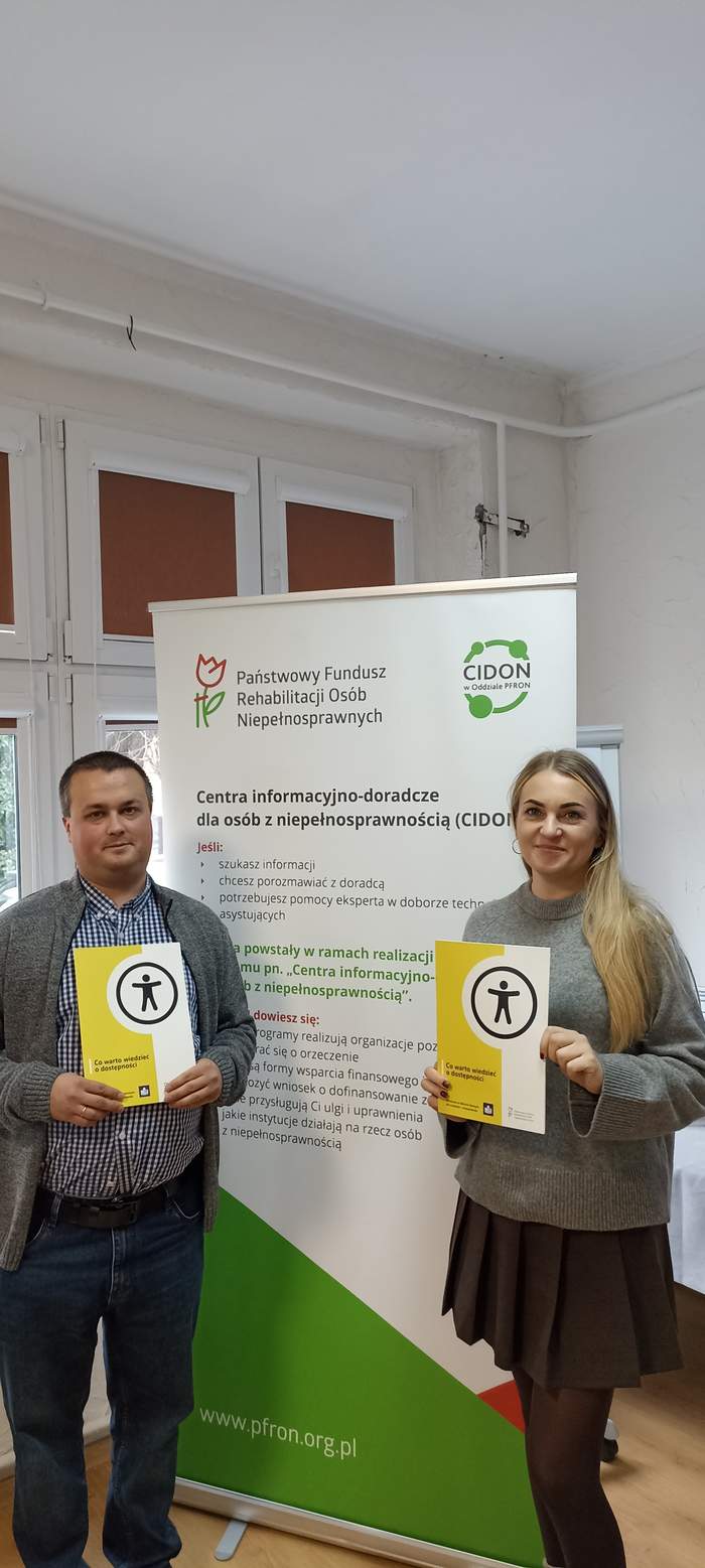 Pokaż zdjęcie: Pracownik PFRON (mężyczna) i pracownik PUP (kobieta) stoją obok siebie w pomieszczeniu Urzędu na tle baneru CIDON i trzymają w rękach żółte broszury dotyczące dostępności