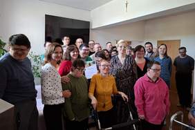 Pokaż zdjęcie: Grupa osób, w tym uczestnicy WTZ, terapeuci i przedstawicielka PFRON, pozuje do wspólnego zdjęcia w sali ośrodka. Na środku widoczna osoba trzymająca dyplom, a wokół niej uśmiechnięci uczestnicy i opiekunowie.