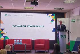 Pokaż zdjęcie: Zdjęcie przedstawia Mariusza Jedynaka, Wiceprezesa Zakładu Ubezpieczeń Społecznych (ZUS), stojącego przy mównicy oznaczonej logotypem ZUS. W tle znajduje się duży ekran z napisem „OTWARCIE KONFERENCJI” oraz kolorowymi geometrycznymi wzorami. Po prawej stronie widoczne są banery ZUS i Państwowego Funduszu Rehabilitacji Osób Niepełnosprawnych. Na scenie ustawione są czerwone fotele oraz stolik z butelkami wody. W dolnej części zdjęcia widać sylwetki uczestników konferencji.