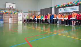 Pokaż zdjęcie: W hali sportowej, pod banerem informującym o XIV Turnieju Sportowo-Rekreacyjnym „Sport dla Wszystkich”, stoją rzędem drużyny reprezentujące różne ośrodki. Uczestnicy ubrani są w kolorowe koszulki, trzymają tabliczki z nazwami placówek. Po lewej stronie kobieta przemawia przy mikrofonie, a nad uczestnikami zawieszono kolorowe balony.