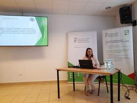 Pokaż zdjęcie: Konferencja, wykład prowadzi kobieta siedząca przy biurku, na którym stoi laptop.