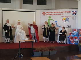 6 osób występuje na scenie, pani z prawej strony siedzi i trzyma na kolanach dużą księgę, pozostali w strojach króla, królowej itp