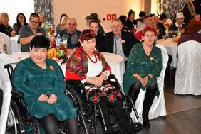 Goście siedzą na sali przy stołach, na pierwszym planie widać trzy kobiety - dwie siedzą na wóżkach, a trzecia na krześłe