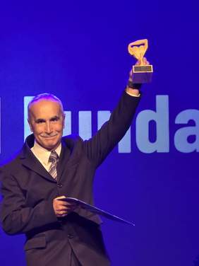 Pokaż zdjęcie: Mężczyzna w garniturze stoi na scenie z niebieskim tłem i trzyma w górze statuetkę. Uśmiecha się, w drugiej ręce trzyma teczkę